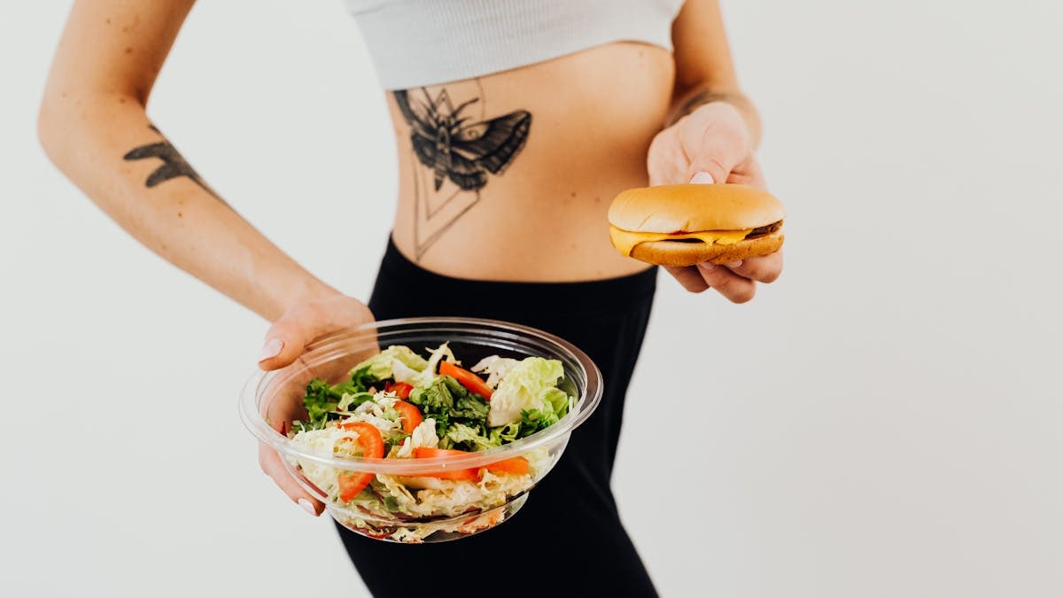 A woman with a tattoo holding a healthy vegetable salad and a cheeseburger, representing food choices.